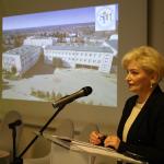 Kobieta przemawia przy mównicy, w tle zdjęcie budynku szkolnego na ekranie.