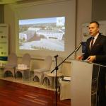 Mężczyzna przemawia przy mównicy na konferencji, w tle prezentacja z budynkiem.