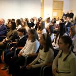 Grupa dzieci i dorosłych siedzi na sali podczas oficjalnego spotkania.