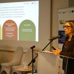 Kobieta w okularach przemawia przy mównicy na tle prezentacji multimedialnej.