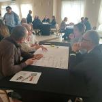 Grupa osób przy stole dyskutuje nad dużą kartką papieru w jasnej sali.