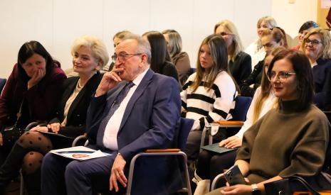 Grupa osób siedzi na krzesłach w sali konferencyjnej, uważnie słuchają.