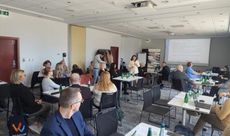 Ludzie siedzący na konferencji, panel dyskusyjny na scenie, ekran z prezentacją.